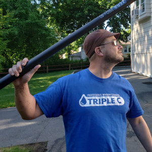 Man in a blue T‑shirt with a TRIPLE C window cleaning logo, carrying a long poles over his shoulder outside a building with trees in the background.