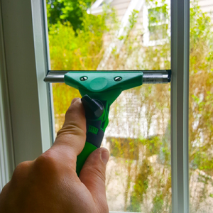 Close-up of a hand gripping a green and black window squeegee, cleaning a glass window.