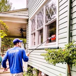 Man in a blue shirt and cap paints exterior window trim of a green house with a long-handled roller.